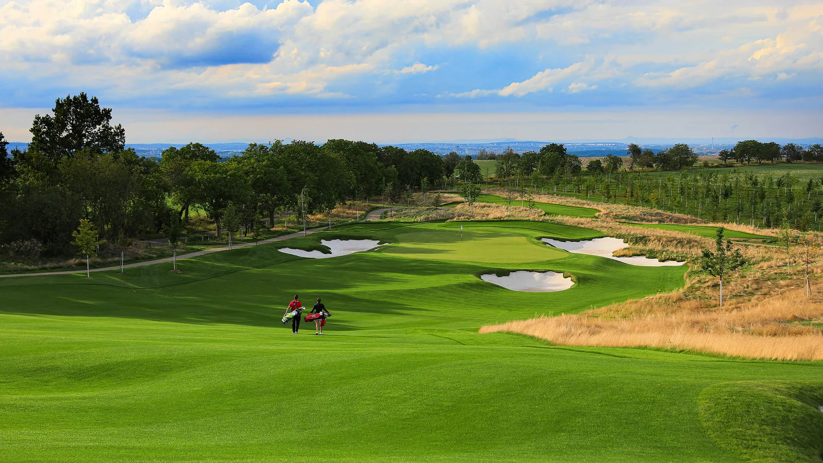 Two golfers walking a broad fairway towards a heavily bunkered green at The Oaks, PGA National Czech Republic